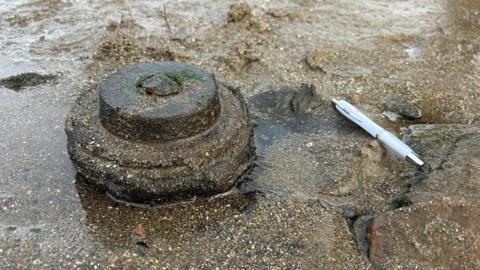 The item found on the beach in Maryport, next to a pen, for scale. The item appears to be made of round sections progressively getting smaller towards the top. The item is a brown colour very close to the colour of the sand around it.