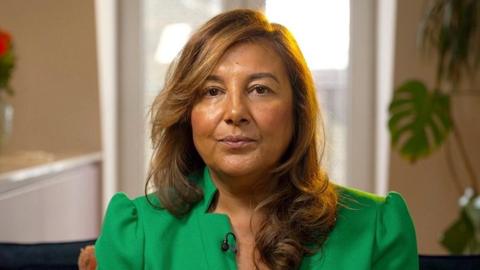 A woman with long brown hair wearing a green dress looks directly at the camera, seated in a brightly lit living space with a large window and green plant in the background.