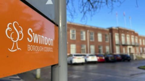 An orange Swindon Borough Council sign in the foreground. Out of focus in the background is the town hall and cars parked outside it on a sunny day.