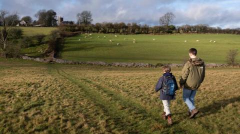 A boy and a young male adult walk through a field in Priddy, Somerset, wearing walking boots and coats. In the background there is a field of sheep, a church and stone walls. 