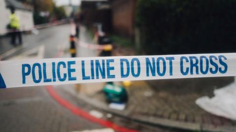 Stock image of a police cordon in place of what looks like a car crash scene with emergency workers and vehicles in the background