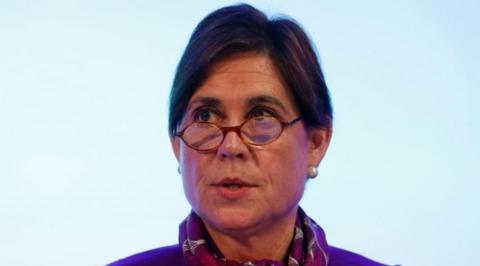 Baroness Kate Lampard, with dark hair, pearl earrings and wearing glasses which are sat halfway down her nose and slightly wonky. She is against a light blue plain background