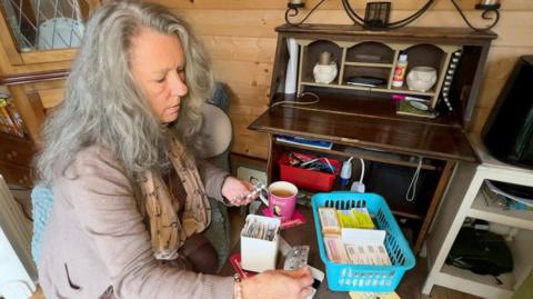 A woman in a beige cardigan with long grey hair sorts through a box of medication at a table. She is looking at a small blister pack of tablets which she is holding in her right hand.