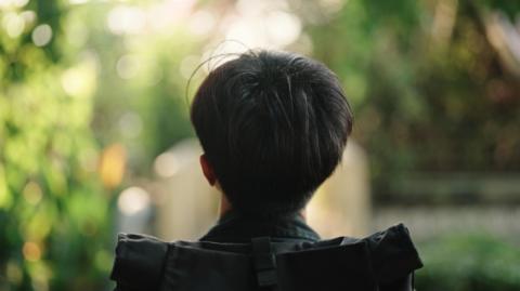 The back of a young man/boy's head. He is either sitting down or walking, but he has a black rucksack on his back.