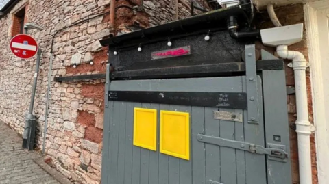 A stone wall with a heavy wooden gate, painted grey. The entrance to a nightclub.