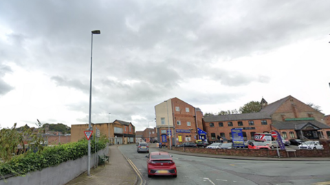 A road junction that bends to the right. Red brick buildings in the background, with a low wall in front of a car park. There is a lamp-post to the left and a sign that reads "give way". 