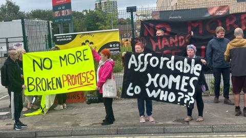 Collyhurst residents hold banners up on a roadside in protest at development proposals for the area. One reads "No more broken promises" on a neon yellow background. The other reads "Affordable homes to who???" on a black background with white lettering.