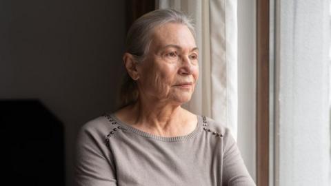 An older woman with long silver hair looks out the bedroom window. She wears a pale grey top with silver beads around the shoulders.