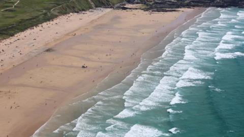 The photo shows some green grass to the top left corner of the picture, which then turns into sand. Towards the middle of the photo the sea can be seen lapping onto the sand with white waves, and to the right of the photo the sea is a dark green/blue colour. 