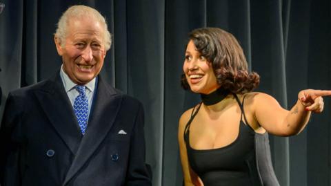 The King smiles on stage next to Raye, who is pointing into the audience