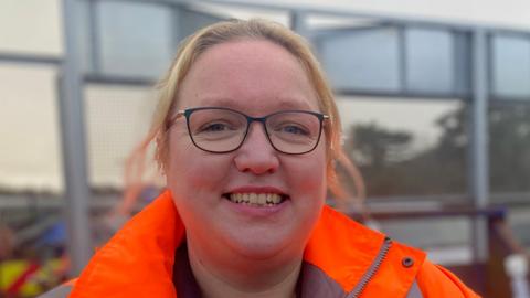 A woman which blonde hair that is tied back smiles at the camera. She is wearing black glasses as well as an orange hi-vis coat.