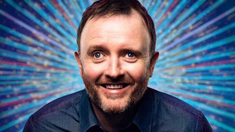 Close-up of Chris McCausland looking into the camera with multi-coloured Strictly branding in the background