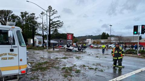 A small white car flipped over in front of a large shop and car park in Scotts Valley. A fire truck and fireman can be seen in the foreground standing alongside debris from nearby trees. 