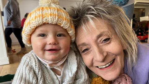 Baby grinning with woolly hat on and an older woman grinning next to him 