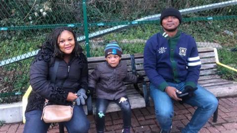 Sue Agazie smiling into the camera while sitting on a park bench next to her son and husband. She has long, dark hair and is wearing a black coat and brown bag.