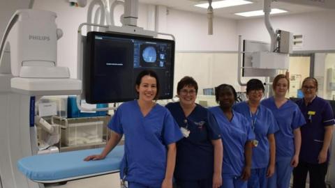 Six hospital staff wearing blue scrubs stand in front of a blue procedure bed, which is next to the new equipment.