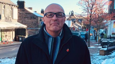 Stocksbridge Towns Deal Board co-chairman Yuri Matischen in large coat with scarf. He has a bald head and black rimmed glasses on.