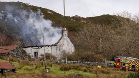 Smoke rises from the ruins of the single-storey, white walled property. There is a fire engine parked just below the property and a small hill in the background.