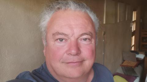 Duncan Ridgley, looking at the camera, he has grey hair, and is wearing a blue T-shirt, he is in a room, with furniture behind. The walls are beige. 