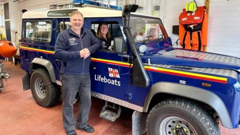 Allan and Helen Thornhill with an RNLI launch land vehicle at RNLI lifeboat station in Flint in north Wales. He is leaning outside it. She is sat in the driving seat 

