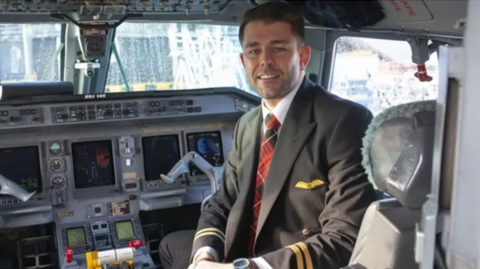 A male pilot in a cockpit facing towards the door and smiling. 