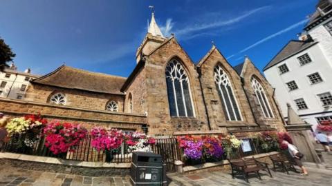 Church with three large stained glass windows, with pink and purple flowers on the black metal fence that runs around the perimeter of the building.