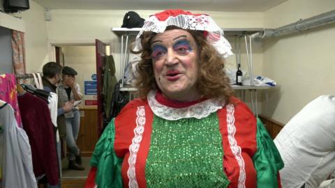 Lloyd Moritmore seen wearing a green and red frilly dress with a long brown curly wig and a white and red hat. He has exaggerated make up with red lips and blue and pink eye shadow.  