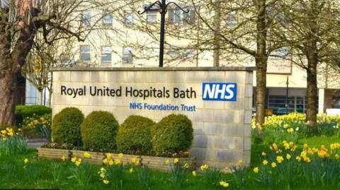 An entrance sign to the Royal United Hospital in Bath bearing the hospital's name and the NHS logo, it is made out of breeze blocks with hedges in front