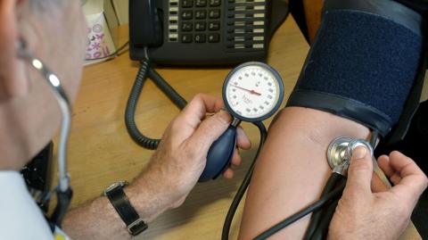 A rear view of a male medical practitioner holding a stethoscope to the inside of person's elbow with his right hand. Above the elbow is a Velcro-covered armband. The medic is holding a hand pump with a pressure meter on top.