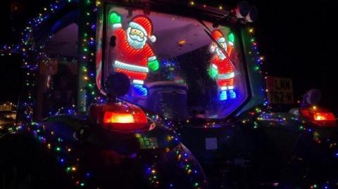 The back window of a tractor, decorated with two light-up Santa Claus figures inside the glass. The rest of the exterior of the tractor is decorated with multicoloured Christmas lights. 