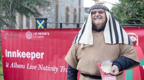 An actor playing the innkeeper stands in front of a red banner which says that he is the innkeeper. He wears a light brown tunic and a black-and-white headdress and is holding star stickers to give to the children