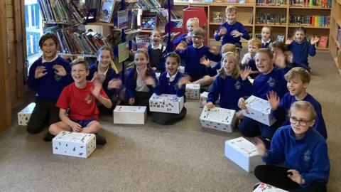 A large group of children aged around 9 - 12 years old smile for a photo. They are sitting as a group on a classroom floor, smiling and waving. Each has a large white shoebox which has been decorated on the outside. Bookshelves can be seen in the background