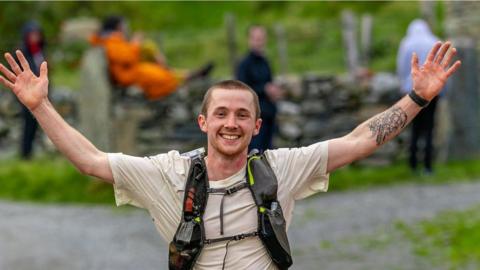 Guy Locke finishing a long distance race with arms-outstretched and smiling at the camera