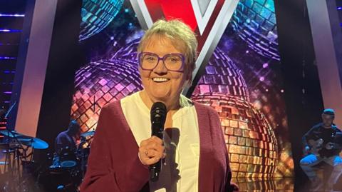 Woman standing on the stage of the Voice UK television programme, holding a microphone and smiling