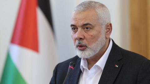 Ismail Haniyeh speaking into a microphone with the Palestinian flag in the background.