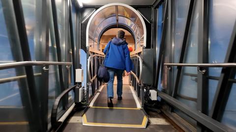 Man in blue jacket, blue jeans and dark boots walks through the glass tunnel on to MV Glen Sannox ferry.