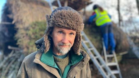 Scot AnSgeulaiche stands in front of a building while another thatcher, wearing hi-vis, works on the roof, halfway up a ladder. He wears a deerstalker hat, a pale green utility jacket, a green fleece and a khaki knitted jumper. 