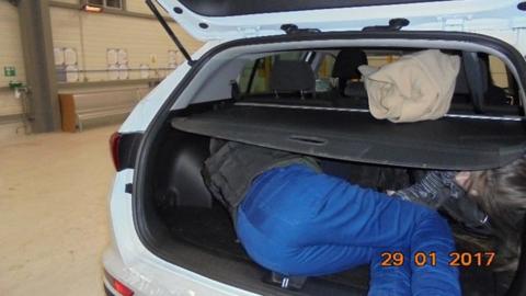 A photograph of two people crammed into the back car of a boot with a time stamp of 29.1.2017. One person is facing outward and lying on their side. A second person in a dark jacket and blue jeans is facing inward, also lying on their side. The parcel shelf is pulled over the top of them.