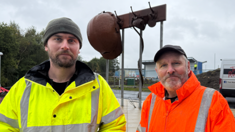 Two men wearing high visibility jackets stand in front of a steel sculpture of a miner's hat hanging from a peg.