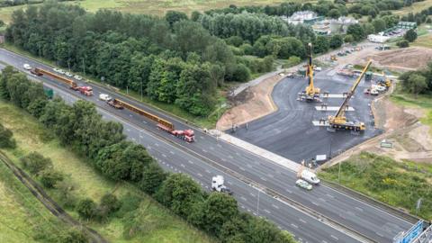 An aerial view picture of the M62 motorway with a work site to its right, with two large cranes. Two lorries can be seen on the motorway carrying two large beams. 