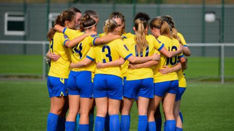 A picture of a group of women footballers in blue and yellow kit huddled on a pitch.