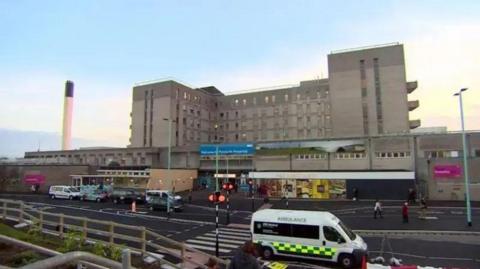 A picture of Derriford Hospital from the car park. There is an ambulance parked on the road. There is a zebra crossing on the road. At the front of the hospital is a Marks & Spencer food hall.