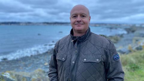 A man in a grey jacket stands near Guernsey's easy coast