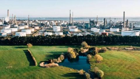 An aerial view of the ExxonMobil Fawley oil refinery shows multiple large white round containers and towers overlooking the Solent, with a field in the foreground and a small pond filled with water and surrounded by trees