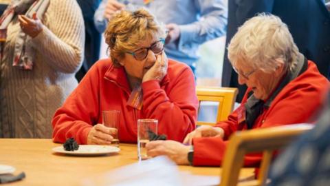 Two women sitting at a wooden table chatting, both are holding glasses of water. Both are wearing red fleeces and glasses, and one has grey hair and the other has greying brown hair. 