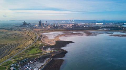 An industrial site on Teesside on the edge of water. There are chimneys bellowing smoke in the distance. 