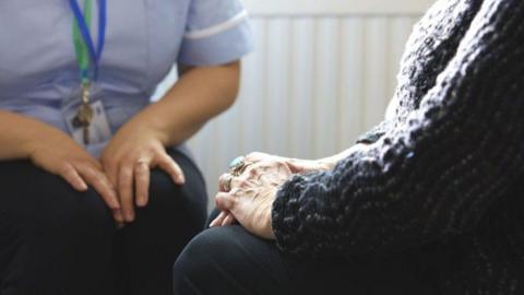 A nurse in a blue uniform sits beside a woman in her home