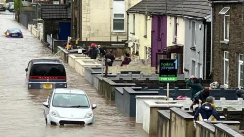 Flooded streets in Sion Street, Pontypridd. Three cars are deep in water in a street that looks more like a river. People wearing raincoats can be seen in the front gardens of terraced houses along the right hand side of the street. There is a pipe over the wall of one of the gardens being used to remove water from a property.