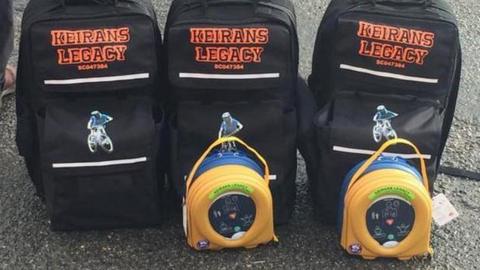 Two round yellow defibrillators in front of three black rucksacks with the words Keiran's Legacy on them and a picture of a mountainbiker.