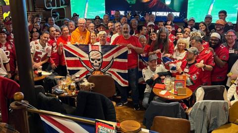 About 50 Buccaneers fans in a bar in London at one of their watch parties. All are crowded together and wearing the team's jersey. Some are holding Great Britain flags, which also have the Buccaneers logo in the middle. 